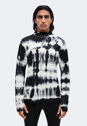 Giovane uomo con capelli scuri fino alle spalle, che indossa un dolcevita tie-dye nero e bianco e pantaloni neri con trama, in piedi davanti a uno sfondo semplice.