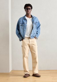 Denim jakna z gumbnimi detajli, bela majica, bež cargo hlače in rjavi patiki. Model stoji z rokama v žepih na lesenem tleh.