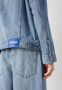 Light blue denim jacket featuring a blue label with "HUGO," silver snap buttons, and a single star accent on the back pocket.