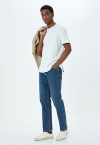 Camiseta blanca de manga corta, camisa de oliva clara caída sobre el hombro, jeans rectos azules y zapatillas blancas. Tela suave, ajuste casual.