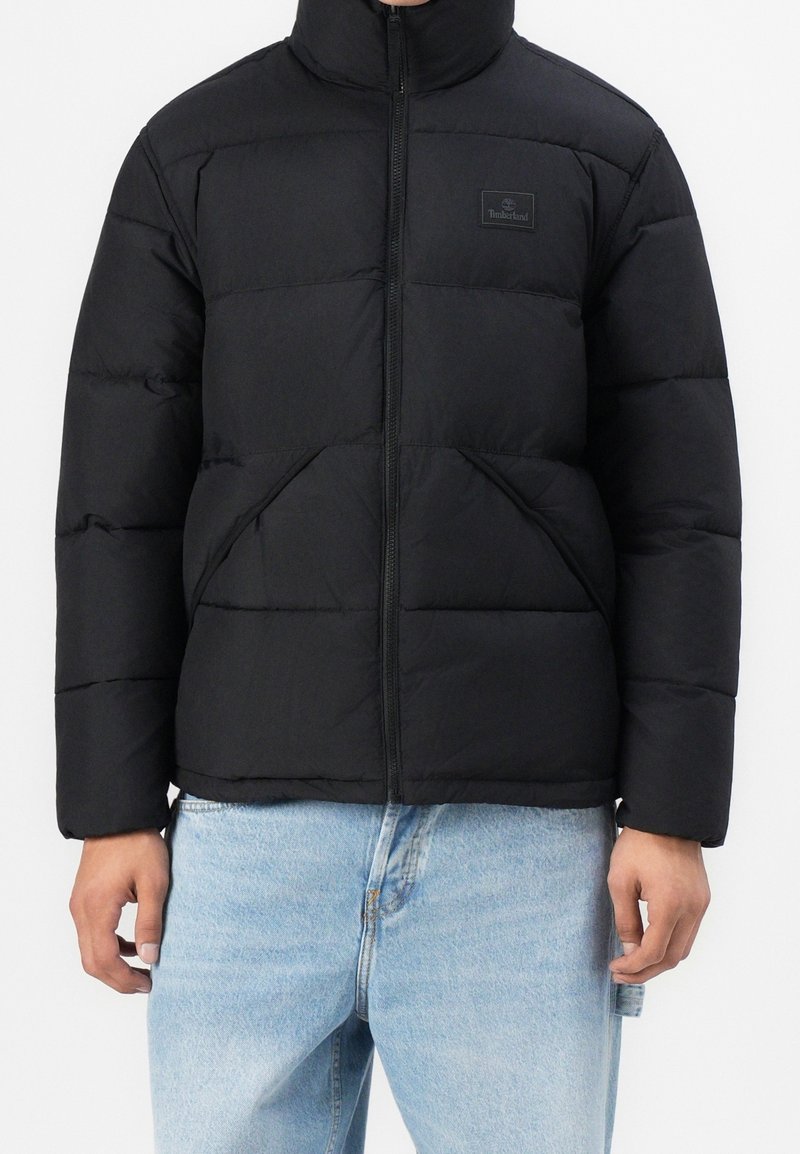 Doudoune noire à col montant, fermeture éclair frontale, poches inclinées, et logo Timberland, portée avec un jean bleu clair.