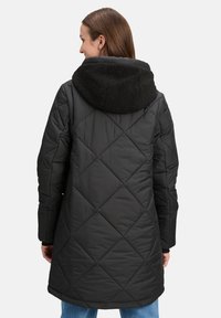 Sort quiltet frakke med en sort shearling-fyldt hætte, lange ærmer og en afrundet kant. Har diagonale syninger og et mellemlangt design.