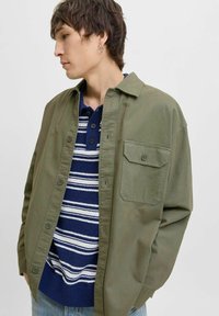 Jeune homme avec des cheveux bruns courts portant une veste verte à boutons sur un t-shirt en tricot rayé bleu et blanc, regardant vers la gauche avec les mains dans les poches.