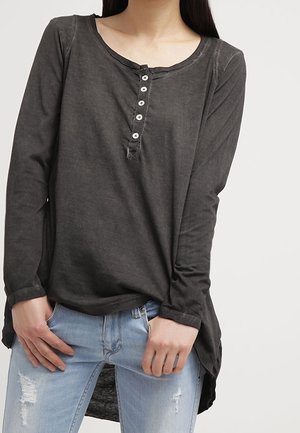 Donkergrijze henley-top met lange mouwen en zes witte knopen, gedragen met lichtblauwe gescheurde spijkerbroek door een persoon met lang donker haar.