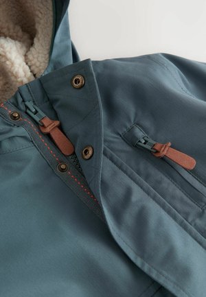 Chaqueta con capucha en azul verdoso que presenta un suave forro crema, cierres de metal, un cierre de cremallera y detalles en cuero marrón. Costuras visibles.