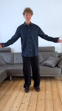 Dark blue button-up shirt with long sleeves, black pants, and black shoes, standing on wooden floor in a living area with a sofa.