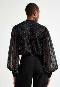 Sort gennemsigtig bluse med puffede ærmer, guldmetal prikmønster og blød tekstur. Sammen med højhalsede sorte bukser.