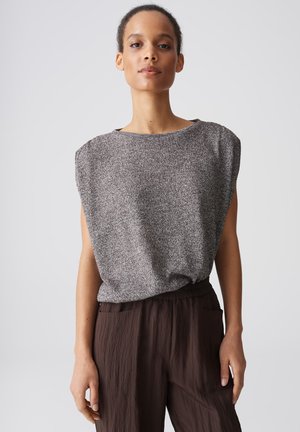 Femme portant un haut en maille gris sans manches avec des épaules rembourrées et un pantalon ample marron foncé, debout devant un fond clair uni.