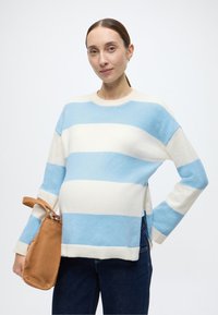 Svītrains džemperis gaiši zilā un krēmkrāsā, izgatavots no mīksta auduma. Tam ir apaļš kakls, brīvs piegriezums un sānu šķēlumi. Kombinēts ar bēša krāsas somu.