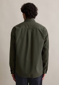 Uomo con capelli ricci che indossa una camicia verde oliva a maniche lunghe, visto da dietro su uno sfondo beige semplice.