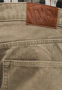 Καφέ δερμάτινο ταμπελάκι Vans στη μέση μπεζ τζιν παντελονιού με ορατή ραφή στην πίσω τσέπη.