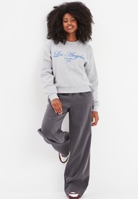 Felpa grigia con scritta blu in corsivo "Los Angeles Creeks 1974" e pantaloni larghi grigi chiari. Il modello si posa in modo casual contro uno sfondo bianco.