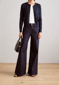 Námořnický cardigan s knoflíky, bílý top, tmavé široké džíny a černá kabelka. Sandály odhalují prsty na nohou; pozadí dřevěné podlahy.