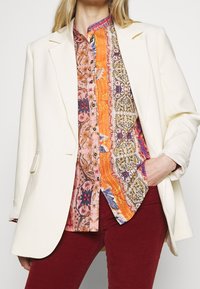 Blazer blanc porté sur une chemise à motif coloré comprenant des nuances d'orange, de rose et de bleu, complété par un pantalon rouge, mettant en avant des textures variées.