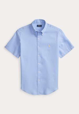 Lys blå kortermet skjorte med knappelukking og krage, med liten gul brodert logo på brystet, lagt flatt på hvit bakgrunn.