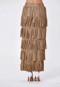 Jupe à franges en simili cuir beige, composée de plusieurs couches de longues franges. La ceinture est élastique, et la jupe tombe jusqu'aux chevilles.