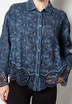 Kobieta ubrana w niebieską bluzkę z długim rękawem z teksturowanym kwiatowym wzorem i okrągłą koronkową lamówką przy dolnej krawędzi i mankietach, w połączeniu z czarnymi spodniami.