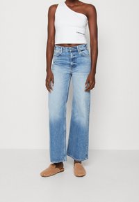 Weißes einseitiges Crop-Top, blaue hochgeschnittene, weite Jeans mit leichtem Verblassen und sandfarbene, kuschelige Slide-Sandalen. Einfaches Design, lässiger Look.