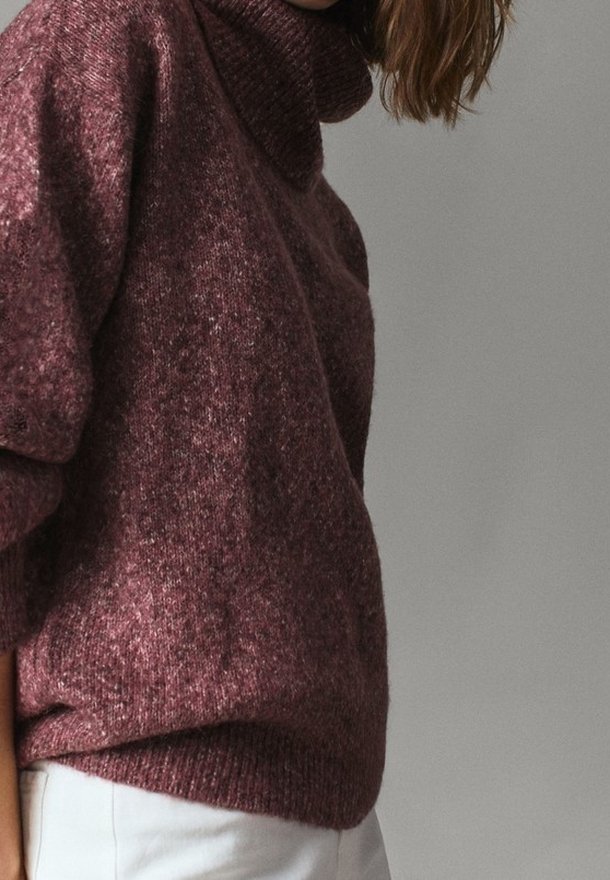 Personne portant un pull à col roulé grenat texturé à manches longues et un pantalon blanc sur un fond gris uni.