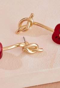 Náušnice v zlatej farbe s dizajnom zapletaného uzla, doplnené červeným kvetom a hladkým, lesklým povrchom. Zapínanie na puzetu.