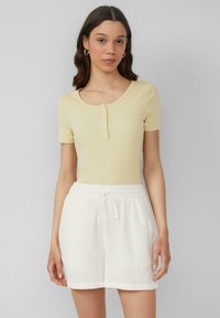 Jasnożółta, prążkowana bluzka z krótkim rękawem i okrągłym dekoltem, z zapięciem na guziki, zestawiona z białymi szortami na sznurki, wykonanymi z teksturowanej tkaniny.