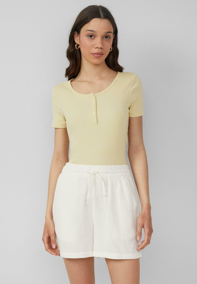 Jasnożółta, prążkowana bluzka z krótkim rękawem i okrągłym dekoltem, z zapięciem na guziki, zestawiona z białymi szortami na sznurki, wykonanymi z teksturowanej tkaniny.