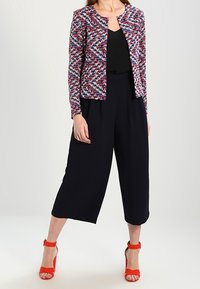 Nainen, joka on pukeutunut moniväriseen kuviolliseen takkiin, mustaan hihattomaan puseroon, leveisiin mustiin cropped-housuihin ja punaisiin korkokenkiin.