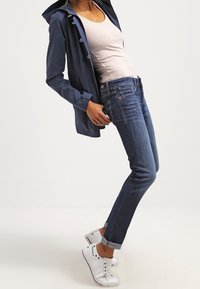 Marinblå vattentät jacka, ljus linne topp, mörkblå jeans, vita sneakers. Jackan har knappar och jeansen är uppvikta vid anklarna.