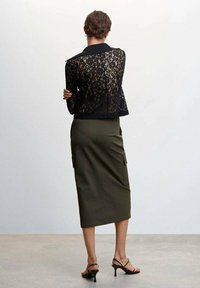 Veste courte en dentelle noire avec un col, associée à une jupe midi vert olive dotée de poches cargo, et des talons noirs à lanières.