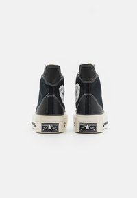 Zapatillas altas negras con suelas de goma crema, costuras blancas y parches de logo redondos. Presentan un tejido texturizado y un diseño distintivo en el talón.