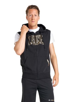 Un homme porte un sweat à capuche noir sans manches et zippé par-dessus un T-shirt blanc, avec un pantalon noir assorti, sur fond blanc.