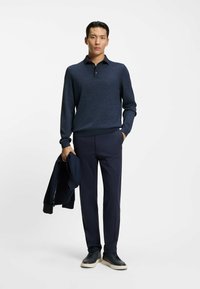 Mörkblå långärmad polo skjorta med en texturerad yta, matchad med raka mörkblå byxor och mörka sneakers. Jacka hålls i högra handen.