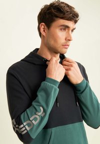 Felpa con cappuccio nera e verde. Composizione in cotone, design a blocchi di colore, con logo argento sulla manica destra. Cappuccio con cordino.