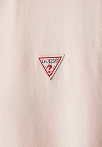 Lys rosa bomullstoff har en liten, trekantet hvit lapp med en rød og svart logo med teksten "GUESS" og et spørsmålstegn.