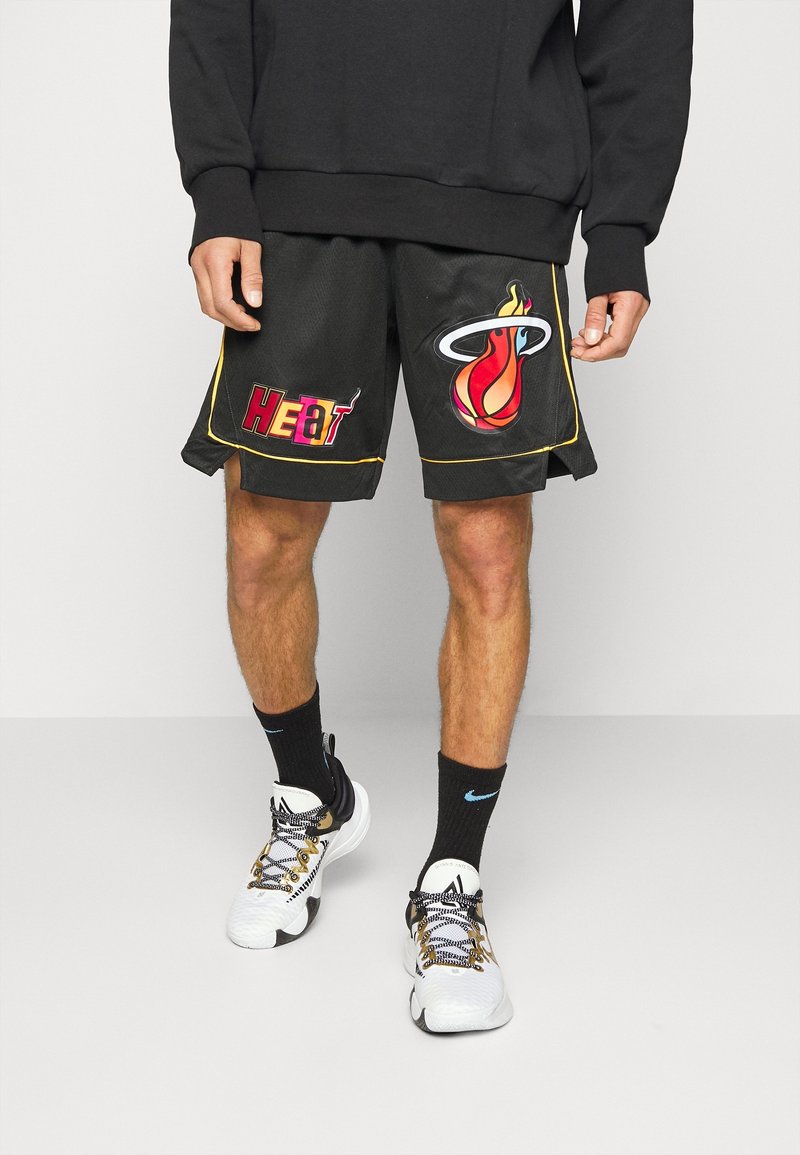Personne portant un short de basketball noir Miami Heat avec logos de flammes et de cerceau, chaussettes noires, baskets blanches et dorées, et un sweat noir.