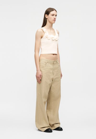 Νεαρή γυναίκα στέκεται πλάγια φορώντας αμάνικη κρεμ κοντή μπλούζα με λουλουδάτες λεπτομέρειες και μπεζ παντελόνες φαρδιές με διακριτικό πουα μοτίβο.