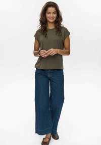 Olivgrünes Kurzarmoberteil, kombiniert mit weit geschnittenen blauen Jeans und dunklen geflochtenen Ballerinas. Das Outfit zeichnet sich durch eine lockere Passform und minimalen Details aus.