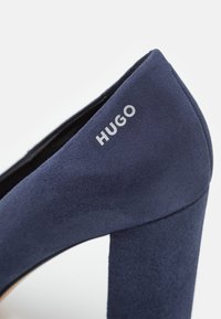 Chaussure à talon haut en daim bleu marine avec talon bloc et logo "HUGO" imprimé en blanc sur le côté extérieur près du col du talon.