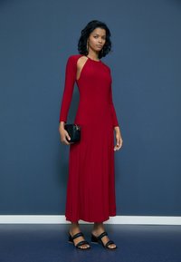 Červené šaty s dlouhým rukávem a výstřihem na rameni, volný silueta, doplněné černou kabelkou a černými pantoflemi na podpatku.