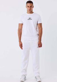 Uomo in piedi di fronte, che indossa una maglietta bianca con logo PXP, pantaloni della tuta bianchi con logo e sneakers bianchi e grigi su sfondo bianco.