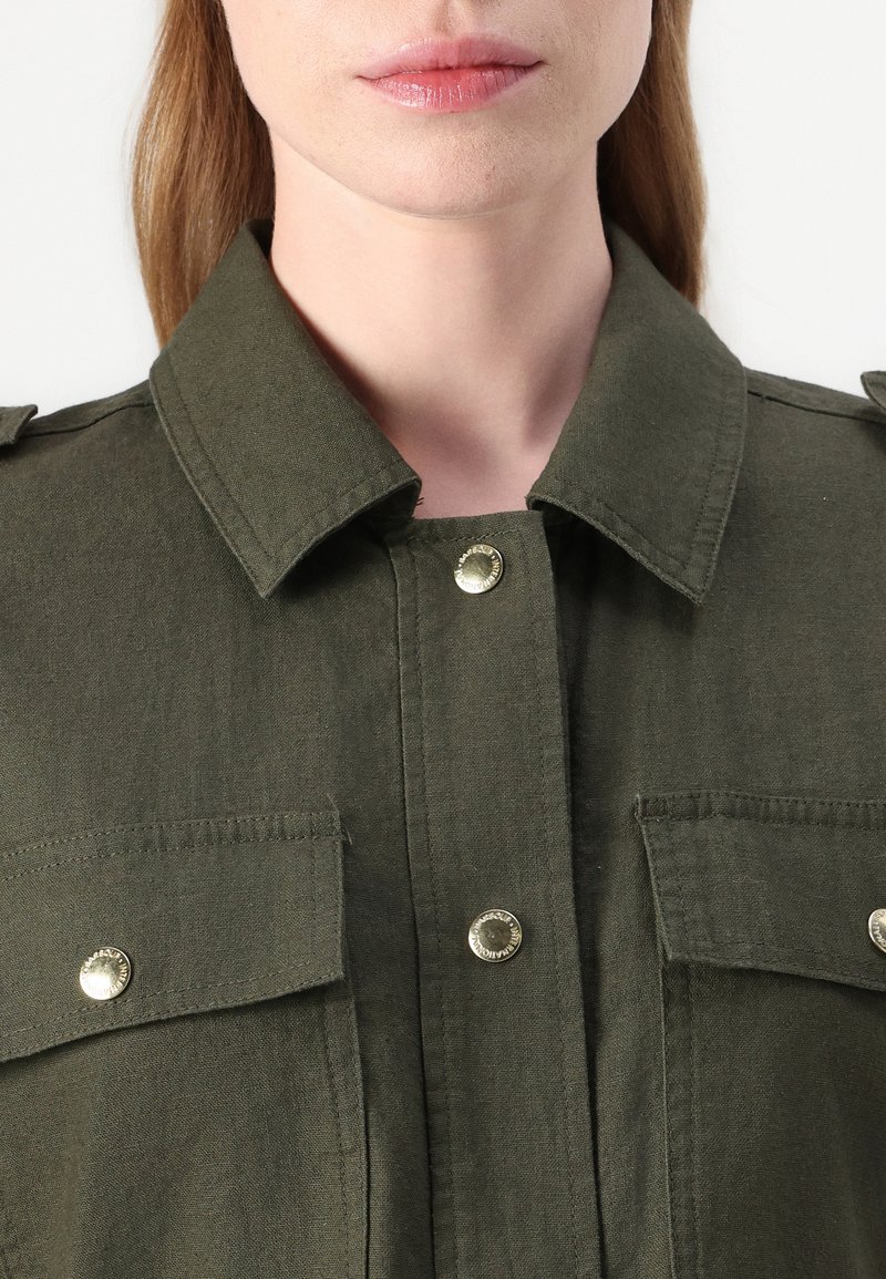 Koszula w oliwkowym kolorze z kołnierzykiem, posiadająca dwie kieszenie na piersi, srebrne zatrzaski oraz pagony na ramionach dla dodatkowego wykończenia.