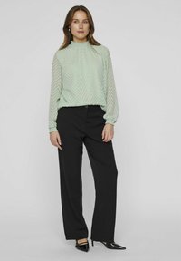 Green patterned blouse with a high neck and long sleeves paired with black wide-leg trousers and pointed black heels.