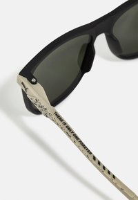 Fekete napszemüveg zöld lencsékkel, matt felülettel, bézs szárakkal, amelyek szöveget és mintákat mutatnak. Fém díszítés a csuklónál.