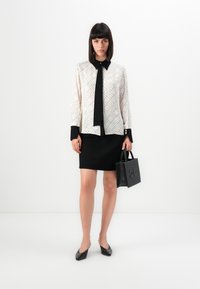 Black skirt paired with a cream blouse featuring a diamond pattern and black accents. The model holds a small black bag with embossed detail.