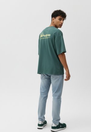 Camiseta de algodón verde con mangas cortas, que presenta un logo gráfico amarillo en la parte trasera. Con pantalones vaqueros azul claro y zapatillas verdes.