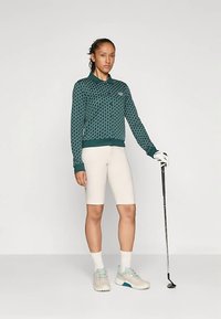 Sweatshirt verde com padrão, gola, calções de ciclismo creme, sapatos claros, segurando um taco de golfe, posando contra um fundo neutro.