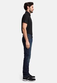En svart kortärmad polo skjorta kombinerad med mörkblå raka jeans och svarta sneakers med vita detaljer. Sidoprofil vy.
