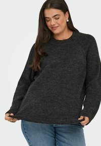 Frau mit langen dunklen Haaren, die silberne Ohrringe, einen dunkelgrauen, gerippten Pullover und blauen Jeans trägt und den Saum des Pullovers hält.