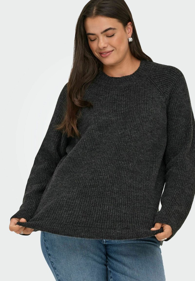 Frau mit langen dunklen Haaren, die silberne Ohrringe, einen dunkelgrauen, gerippten Pullover und blauen Jeans trägt und den Saum des Pullovers hält.