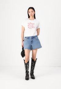 Witte t-shirt met rood grafisch ontwerp, gecombineerd met blauwe denimshorts met rafelige randen en zwarte kniehoge cowboylaarzen met witte stiksels.
